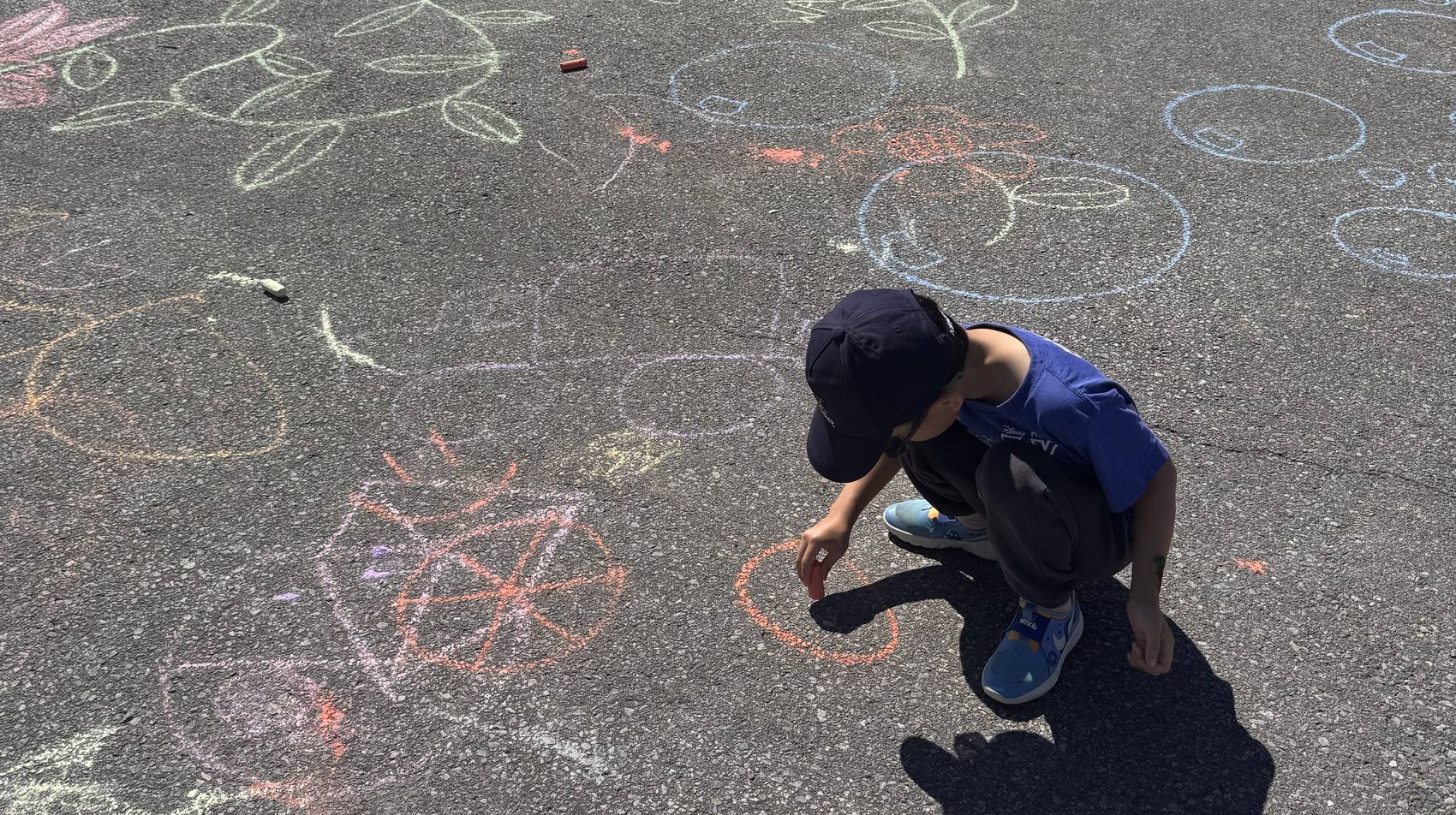 A boy crouched on a sunlit driveway, drawing in orange chalk
